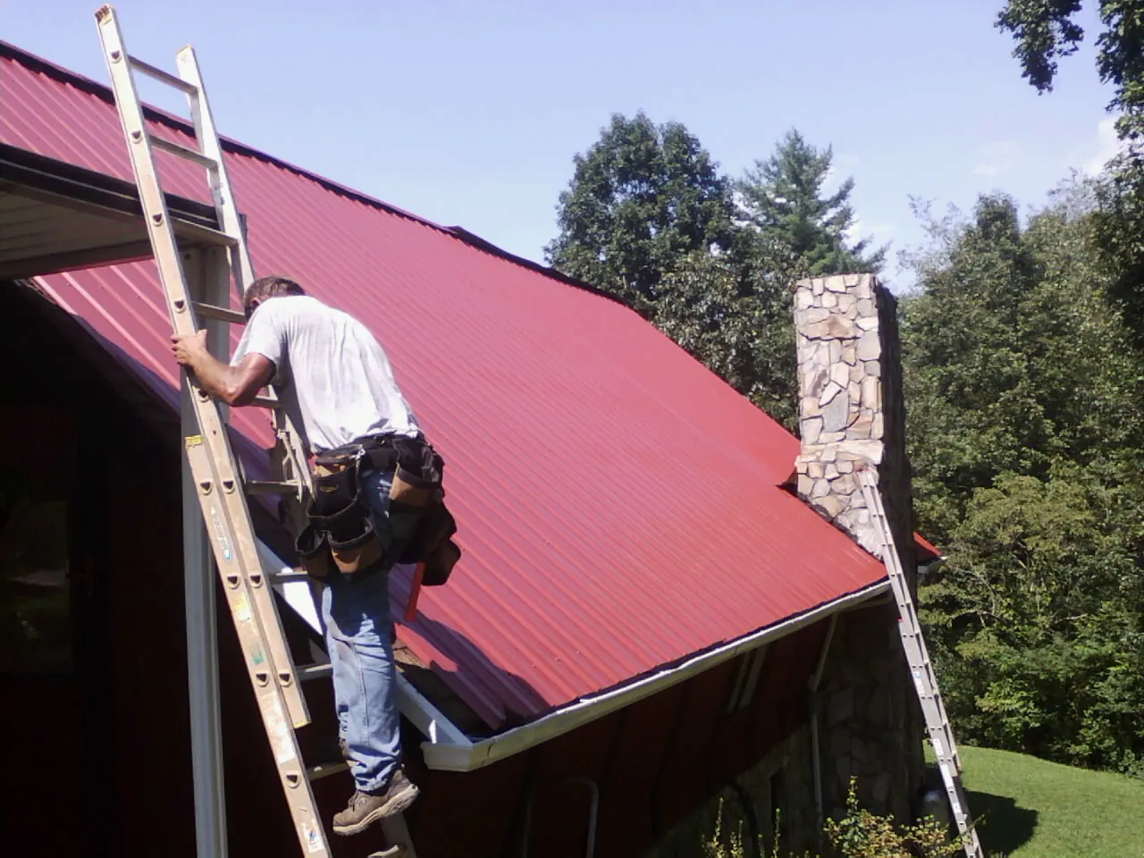 Roofer on a ladder beside a red metal roof and stone chimney — Soffit & Fascia Repair by Roofing Brothers in Yorba Linda